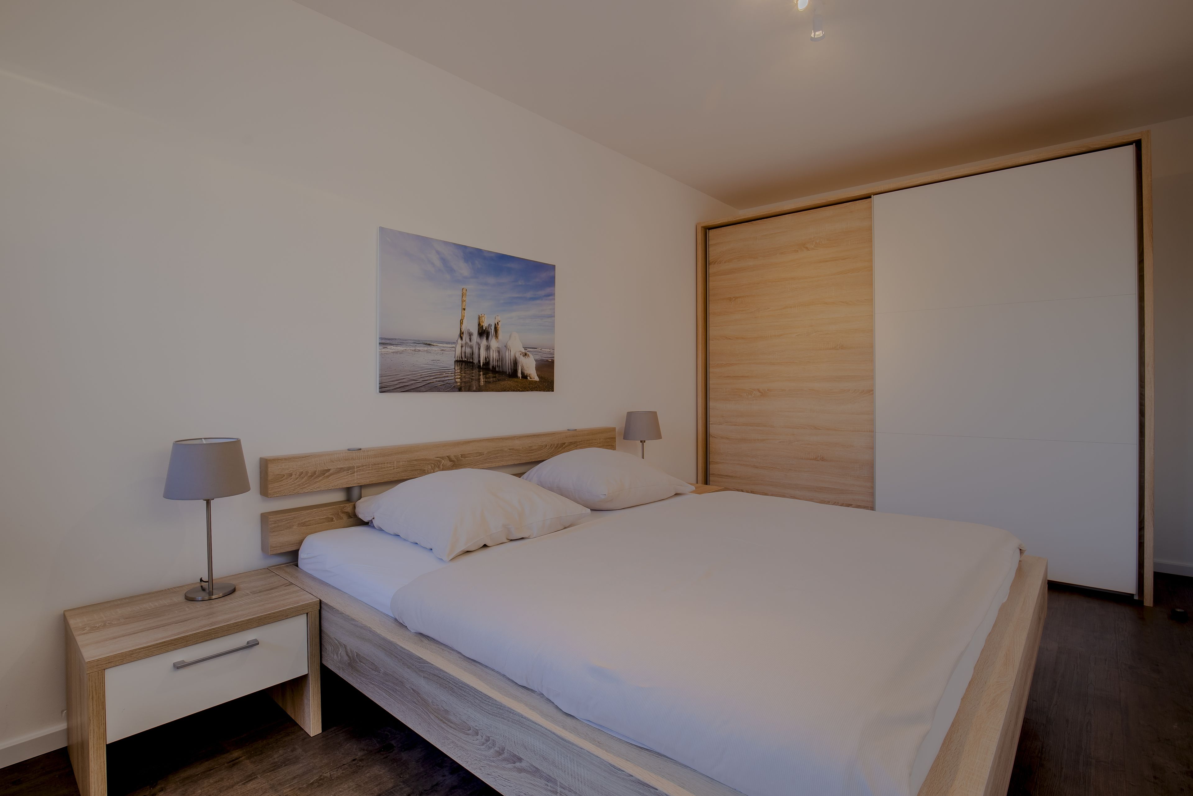A modern bedroom with a wooden headboard, a single bed with white bedding, a nightstand with a lamp, and a wooden wardrobe.