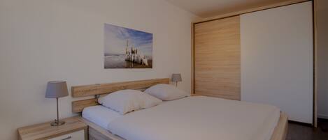 A modern bedroom with a wooden headboard, a single bed with white bedding, a nightstand with a lamp, and a wooden wardrobe.