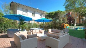 An outdoor patio area with wicker furniture, two blue umbrellas, and a play structure in the background. - Hotel Turquoise (Cesenatico)