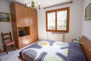 A bedroom with a bed, wooden wardrobe, television, chair, and a window with a view of greenery. - Villa Serena (Portoferraio)