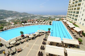 A large swimming pool with multiple lanes, surrounded by sun loungers and umbrellas, with a view of distant hills and buildings. - Goldcity Holiday Homes (Alanya)