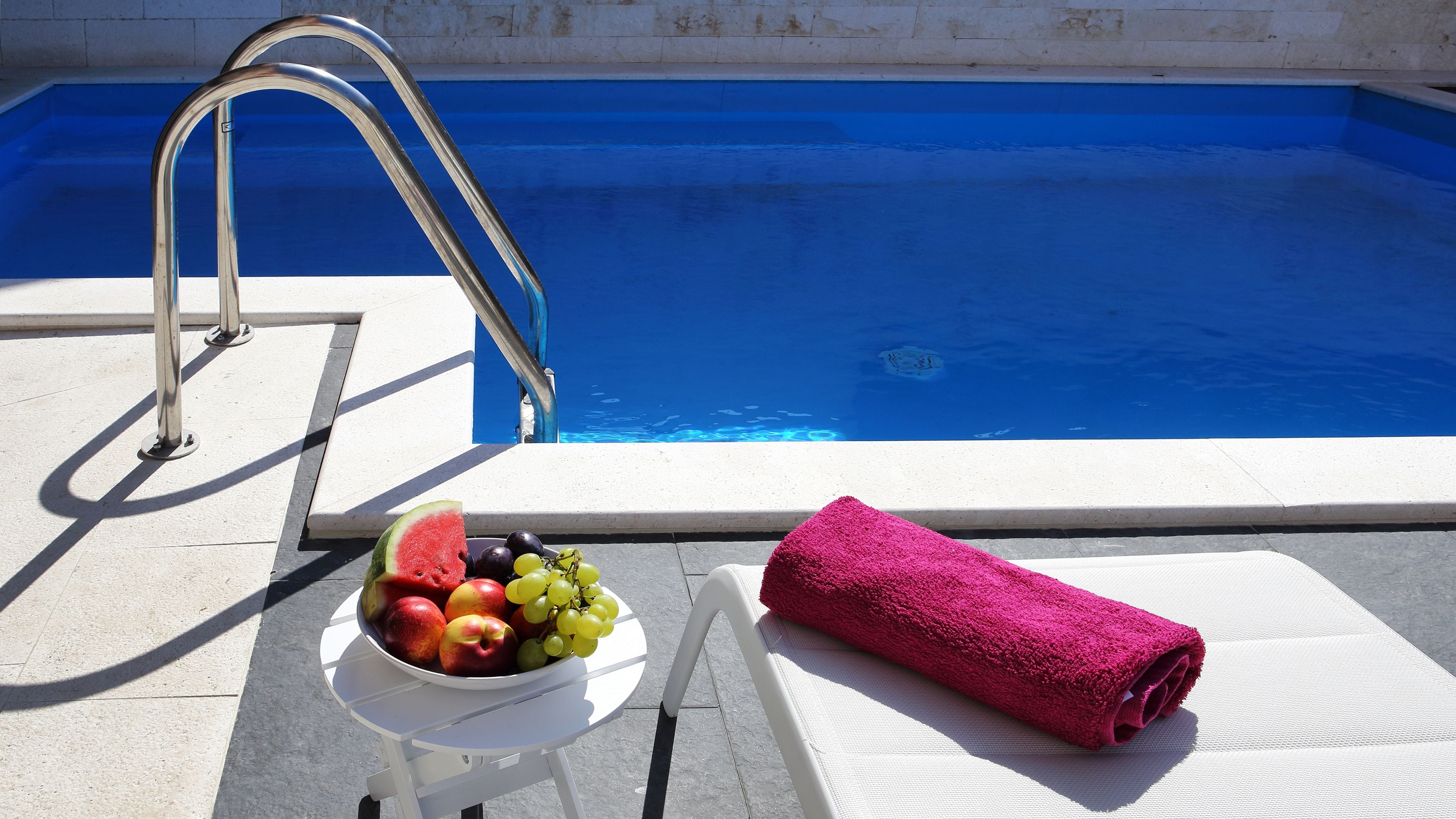 A poolside area with a metal ladder, a bowl of fruit, and a rolled-up towel.