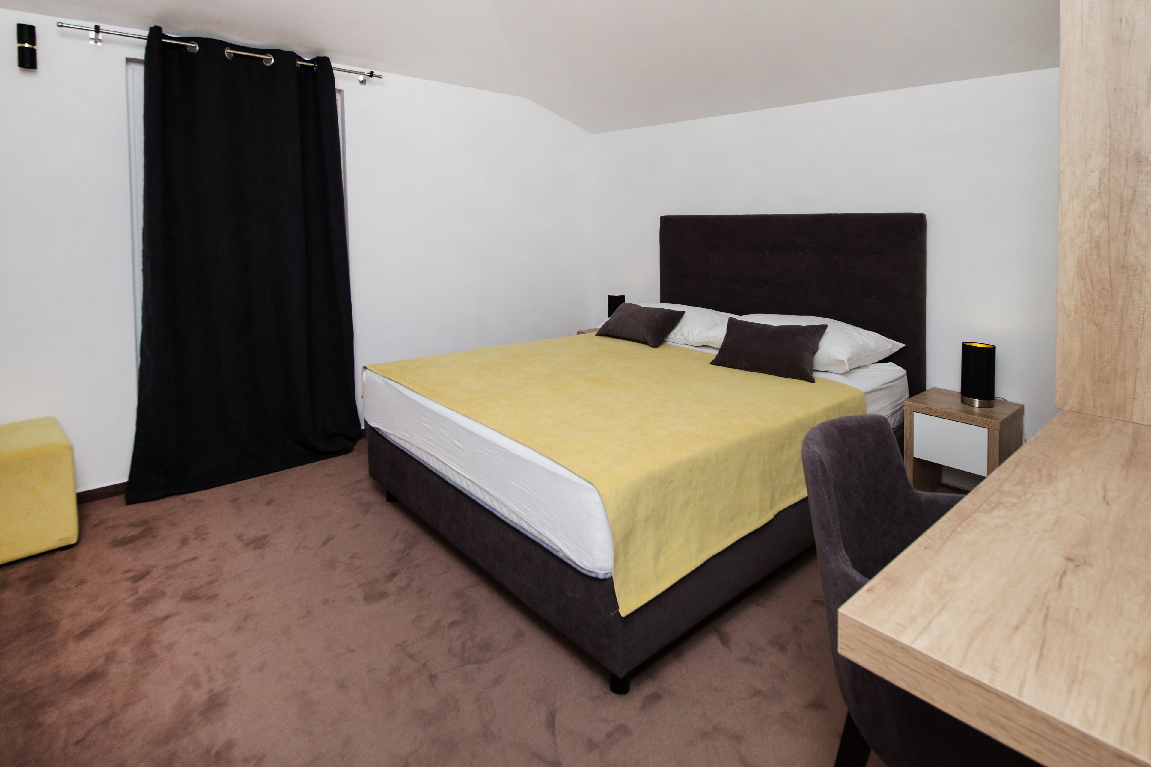A hotel room with a bed, a wooden desk, a chair, a nightstand, and a black curtain.