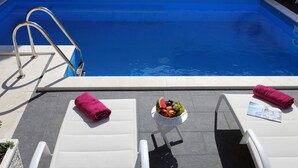 A poolside area with a white lounge chair, a rolled-up towel, a bowl of fruit, and a newspaper. - Villa M (Split)