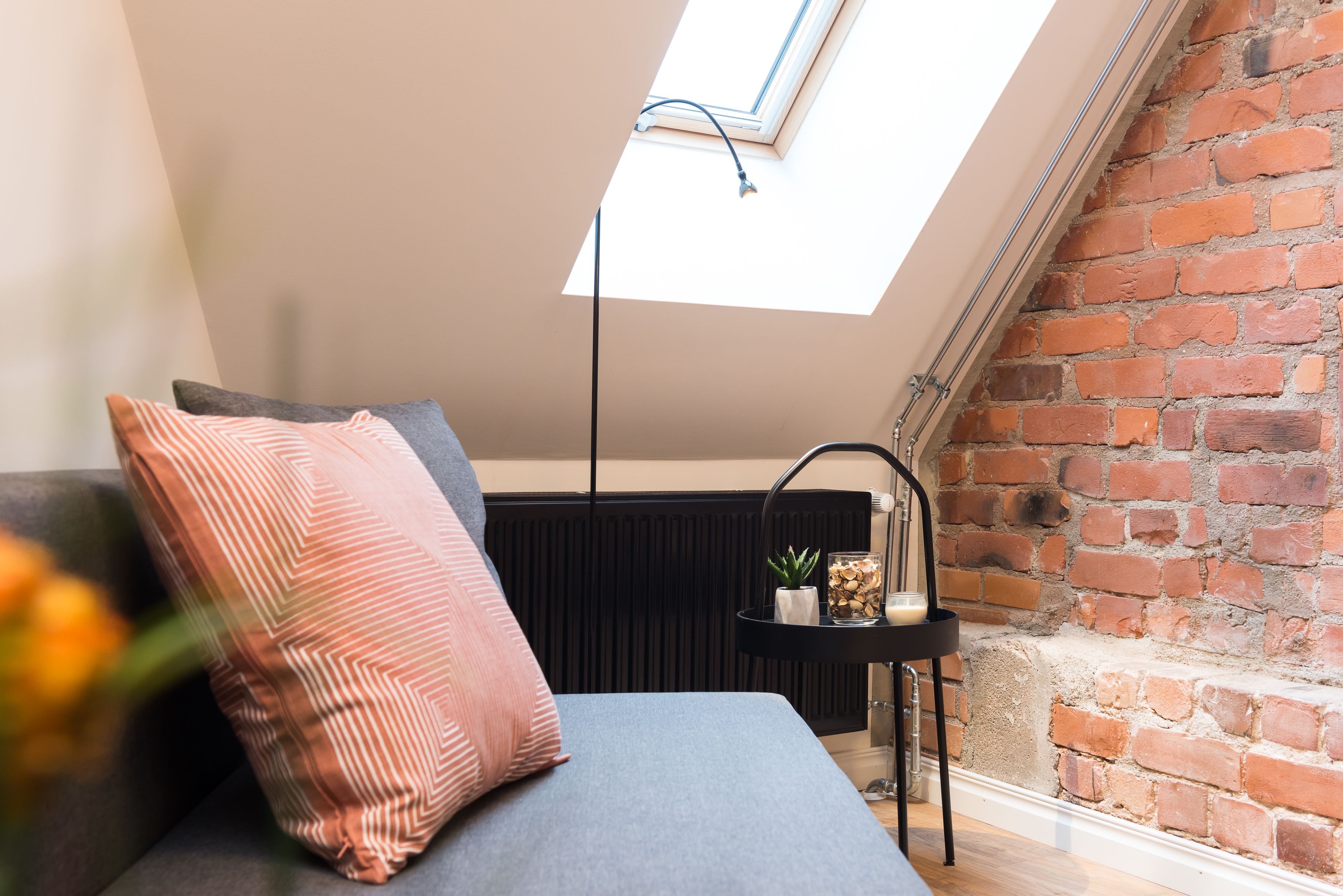 A room with a skylight, a brick wall, a black radiator, a grey sofa, and a small black side table with a plant and candle.