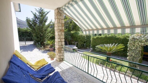 Un balcon avec des fauteuils de jardin bleus et jaunes, un pilier en pierre et une vue sur un jardin où poussent différentes plantes, avec une montagne en arrière-plan.