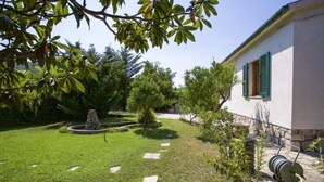 Un jardin bien entretenu, avec un chemin de pierre menant à une fontaine, entouré d’une végétation luxuriante et bordé d’un bâtiment aux volets verts.