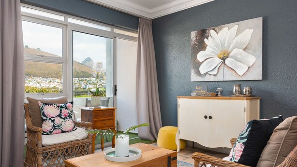 A living room with wicker furniture, a white cabinet, and a large window with a view of a cityscape and mountains. - Sandringham 23 (Cape Town)