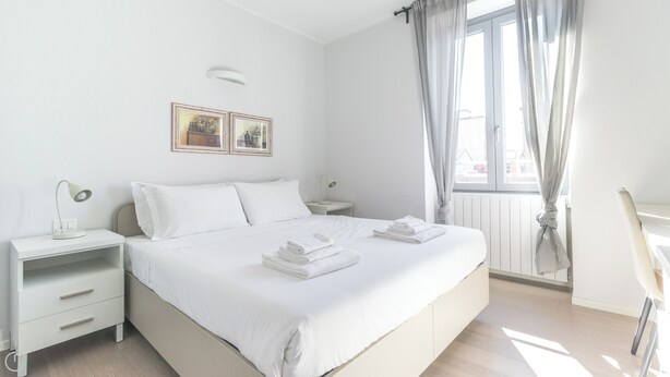 A minimalist bedroom with a bed, two bedside tables, a nightstand, a window with curtains, and a chair. - Italianway Comfort - Bligny 19 B (Milan)