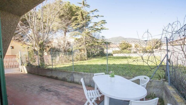 Pátio externo com mesa e cadeiras brancas, cercado por um muro de pedra e uma cerca.