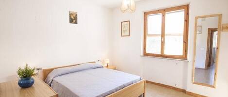 A bedroom with a bed, a bedside table with a lamp, a window, and a mirror.