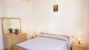 Quarto com cama de madeira, mesa de cabeceira com abajur, cômoda com espelho e duas plantas em vasos.