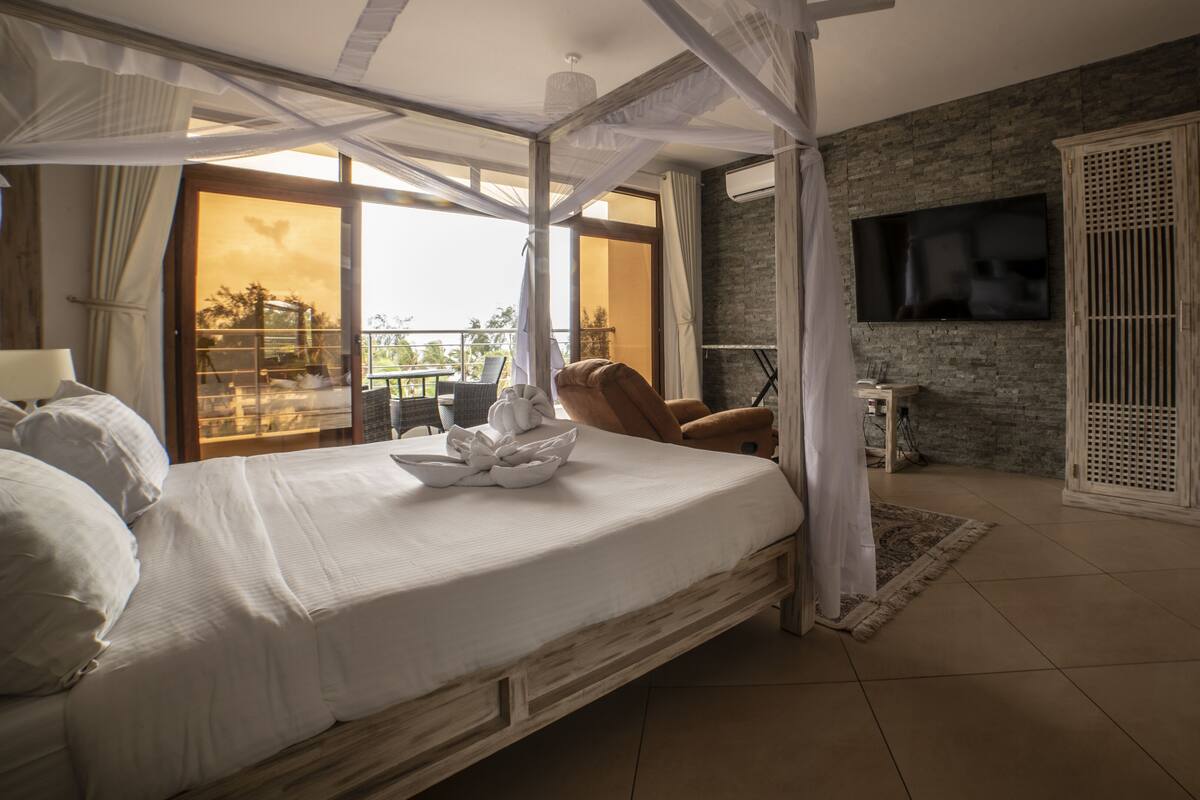 A bedroom with a large bed, a TV, a balcony with a view, and a wooden wardrobe.