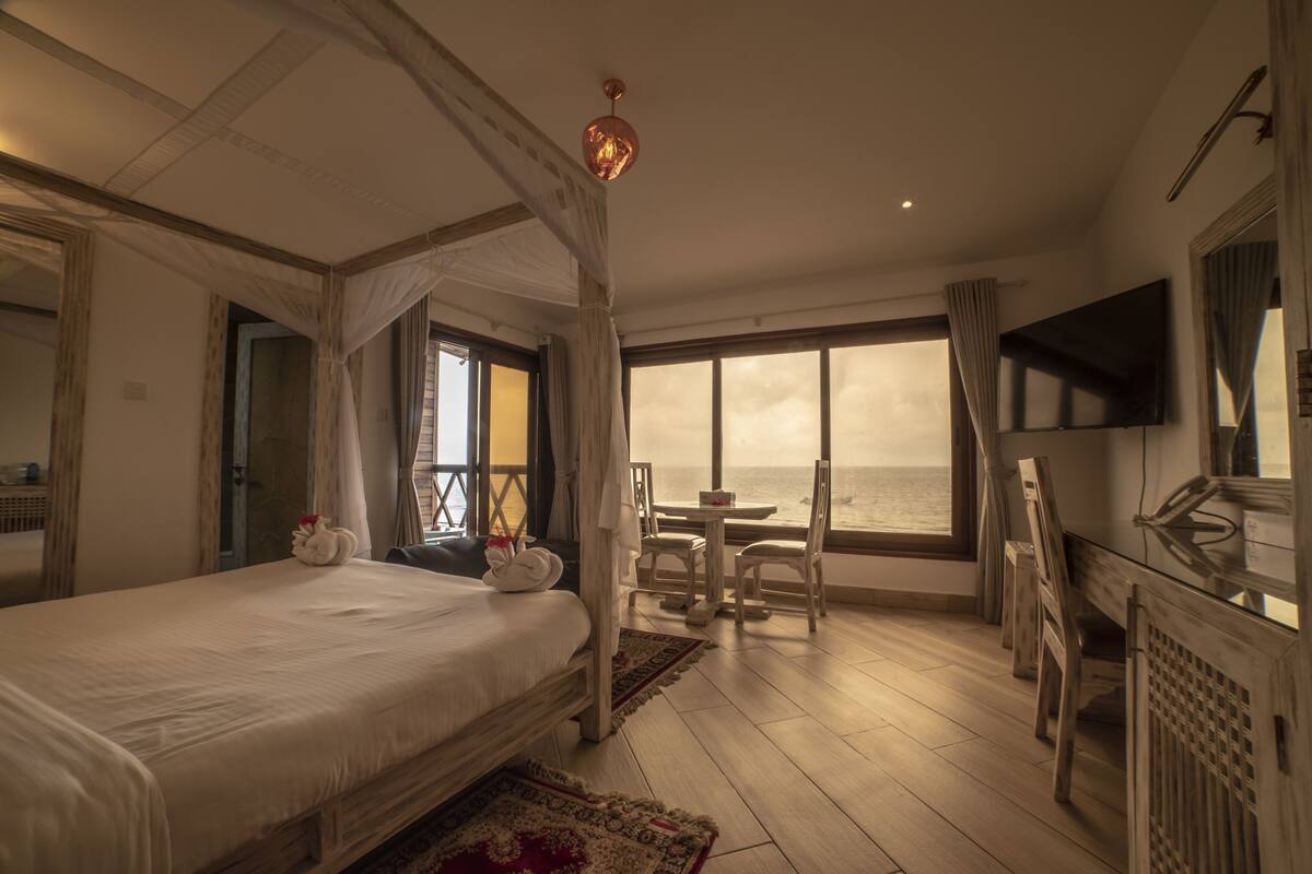 A bedroom with a large bed, a TV, a balcony with a table and chairs, and a view of the sea.