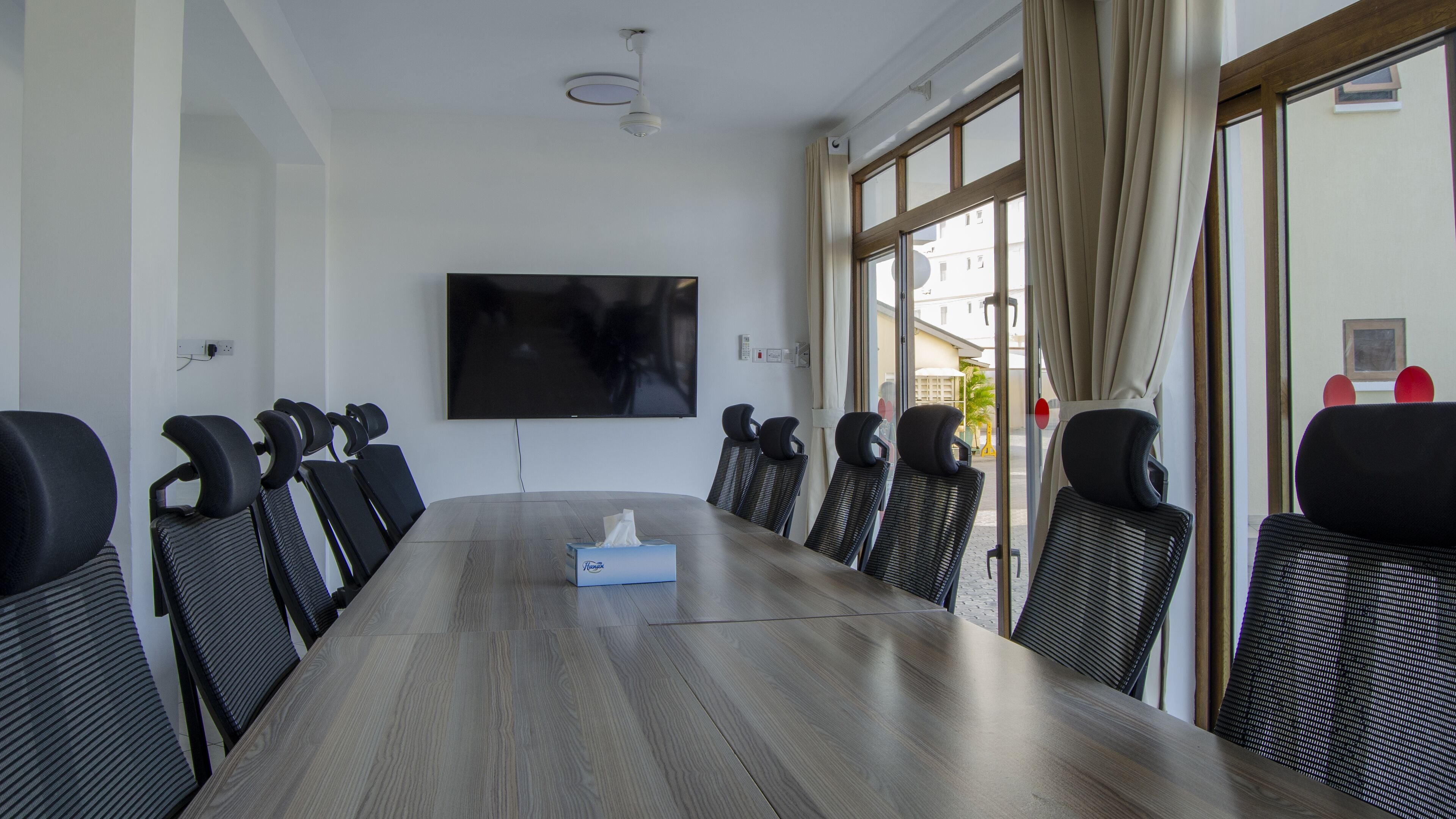 A conference room with a long table, black chairs, a flat-screen TV, and a window with curtains.