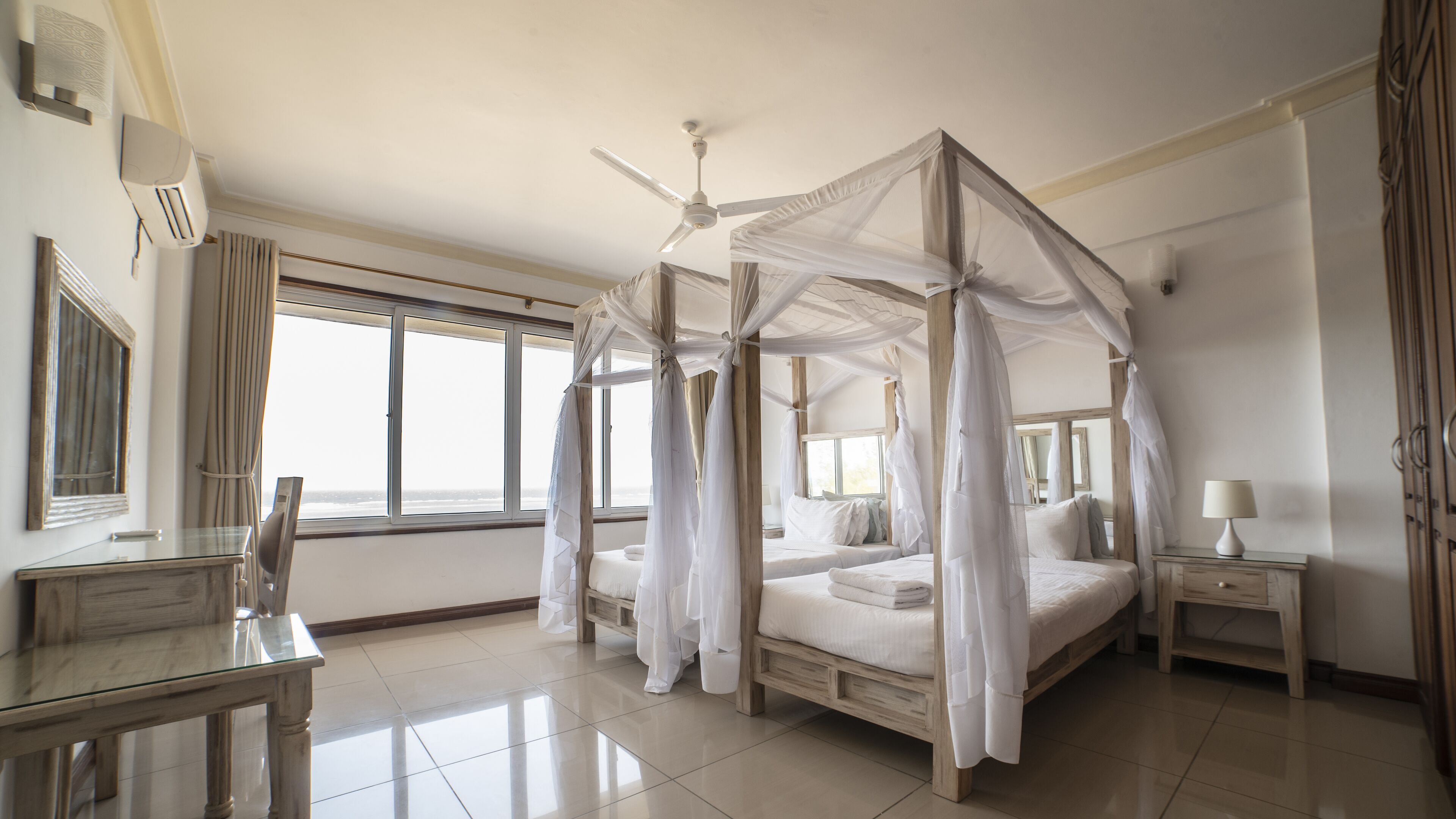 A spacious bedroom with a four-poster canopy bed, a ceiling fan, and a large window providing a view of the beach.