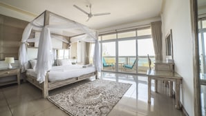 A bedroom with a four-poster bed, a ceiling fan, a large window with a view of the sea, a balcony with a chair, and a nightstand with a lamp.