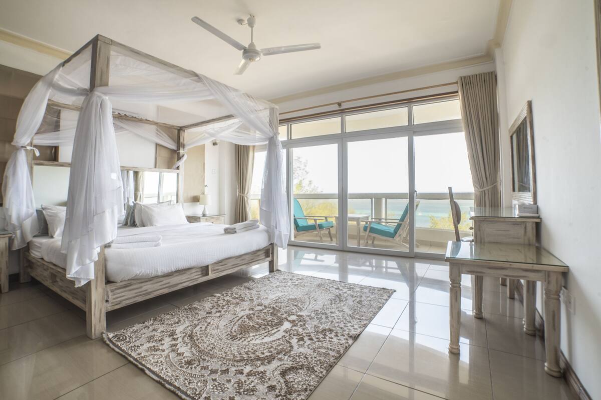 A bedroom with a four-poster bed, a ceiling fan, a large window with a view of the sea, a balcony with a chair, and a nightstand with a lamp.