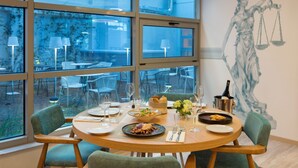 A dining area with a round table set for four, large windows, and a mural of a figure holding scales. - ibis Styles Athens Routes (Athens)