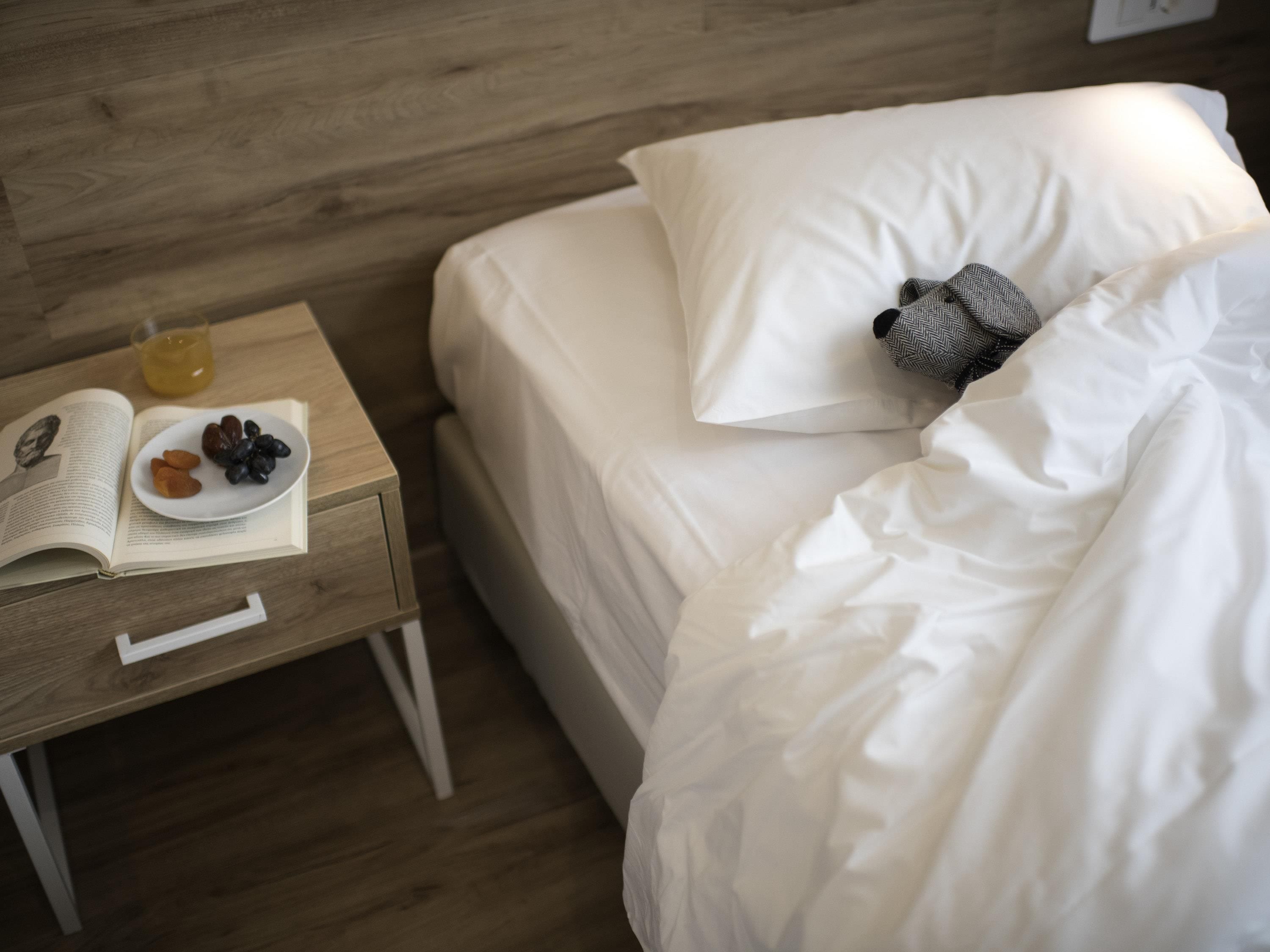 A neatly made bed with white bedding, a pillow, and a small knitted item. A bedside table holds a book, a plate with dates, and a glass of juice.