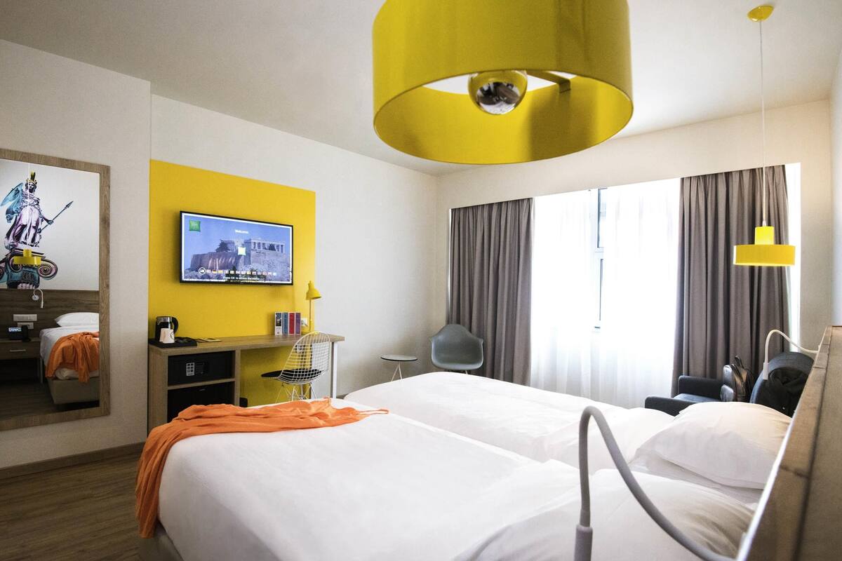 Et hotelværelse med to senge, et fjernsyn på væggen, et skrivebord med stol og et vindue med gardiner.
