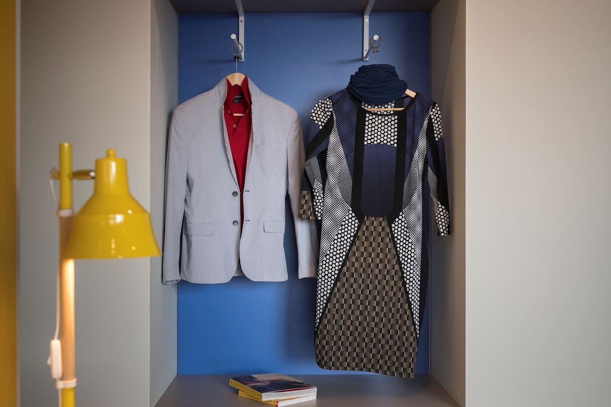 A room with a wooden desk, a yellow desk lamp, a blue wall, a hanging coat rack with two suits, and a pair of shoes on the floor.