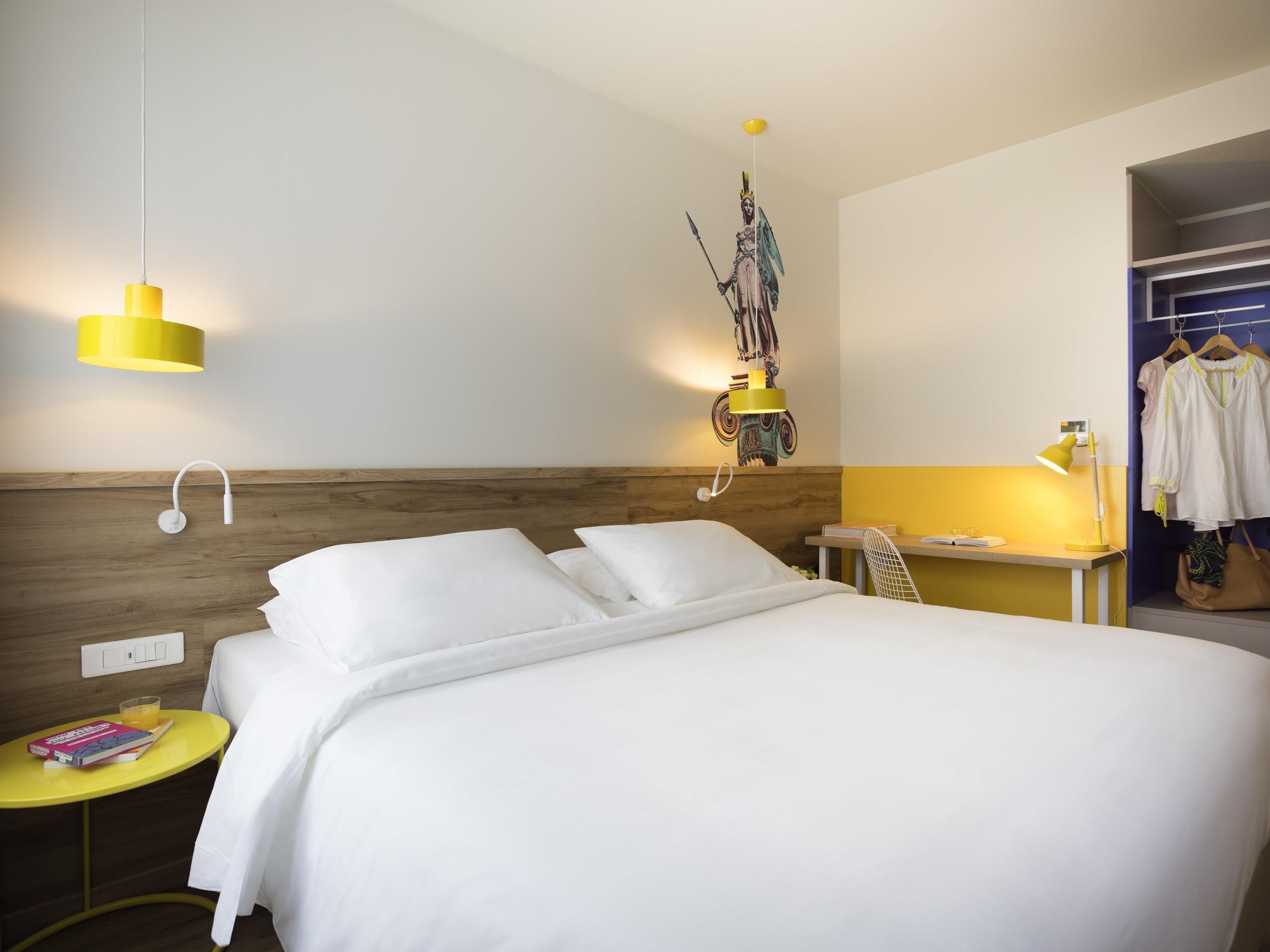 A hotel room with a bed, a small table with a book, a hanging decorative item, and a clothing rack.