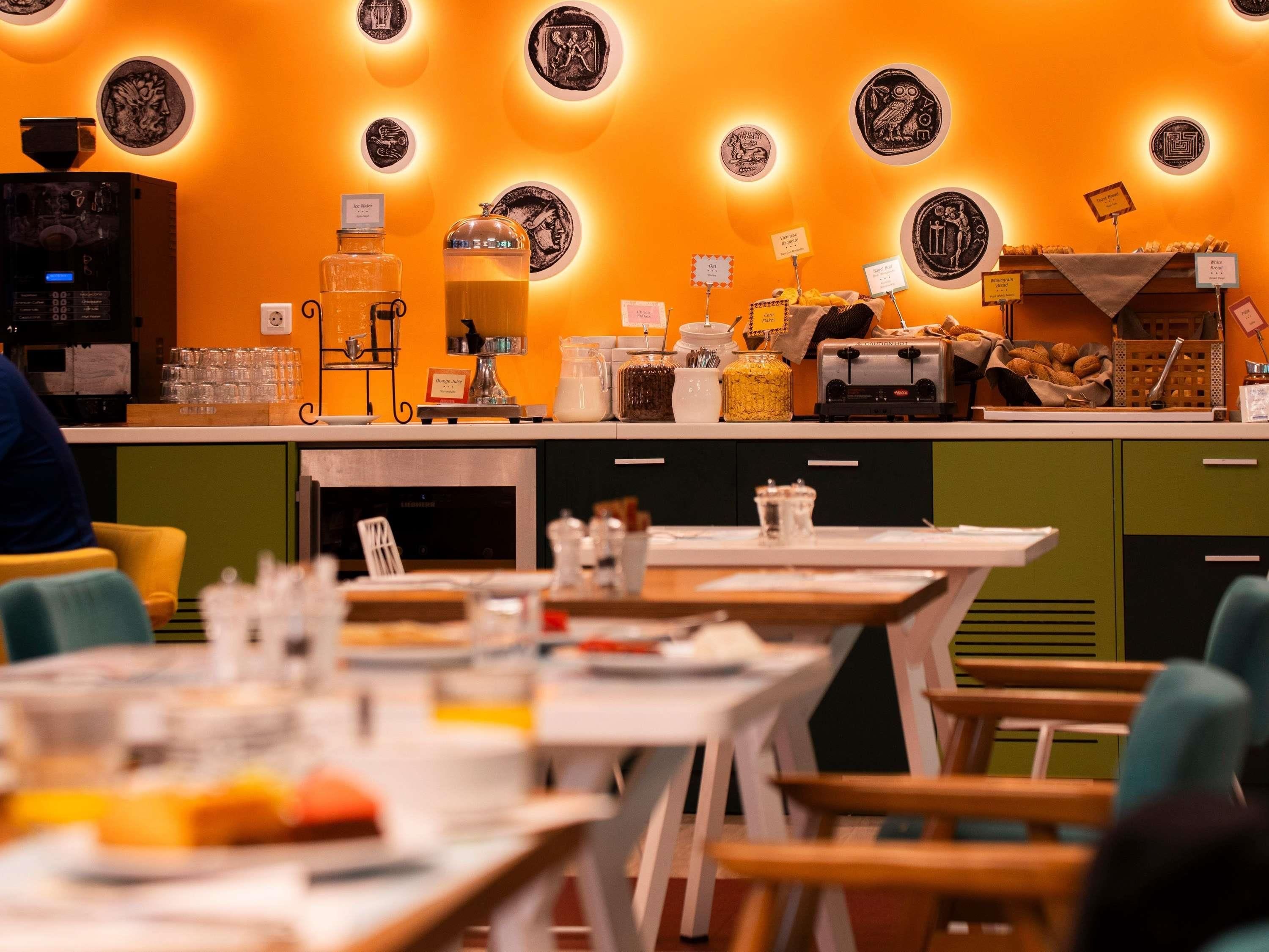 A restaurant interior with a counter displaying food items and a coffee machine. Tables are set with chairs, and the walls are decorated with circular designs.