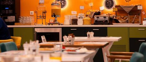 A restaurant interior with a counter displaying food items and a coffee machine. Tables are set with chairs, and the walls are decorated with circular designs.