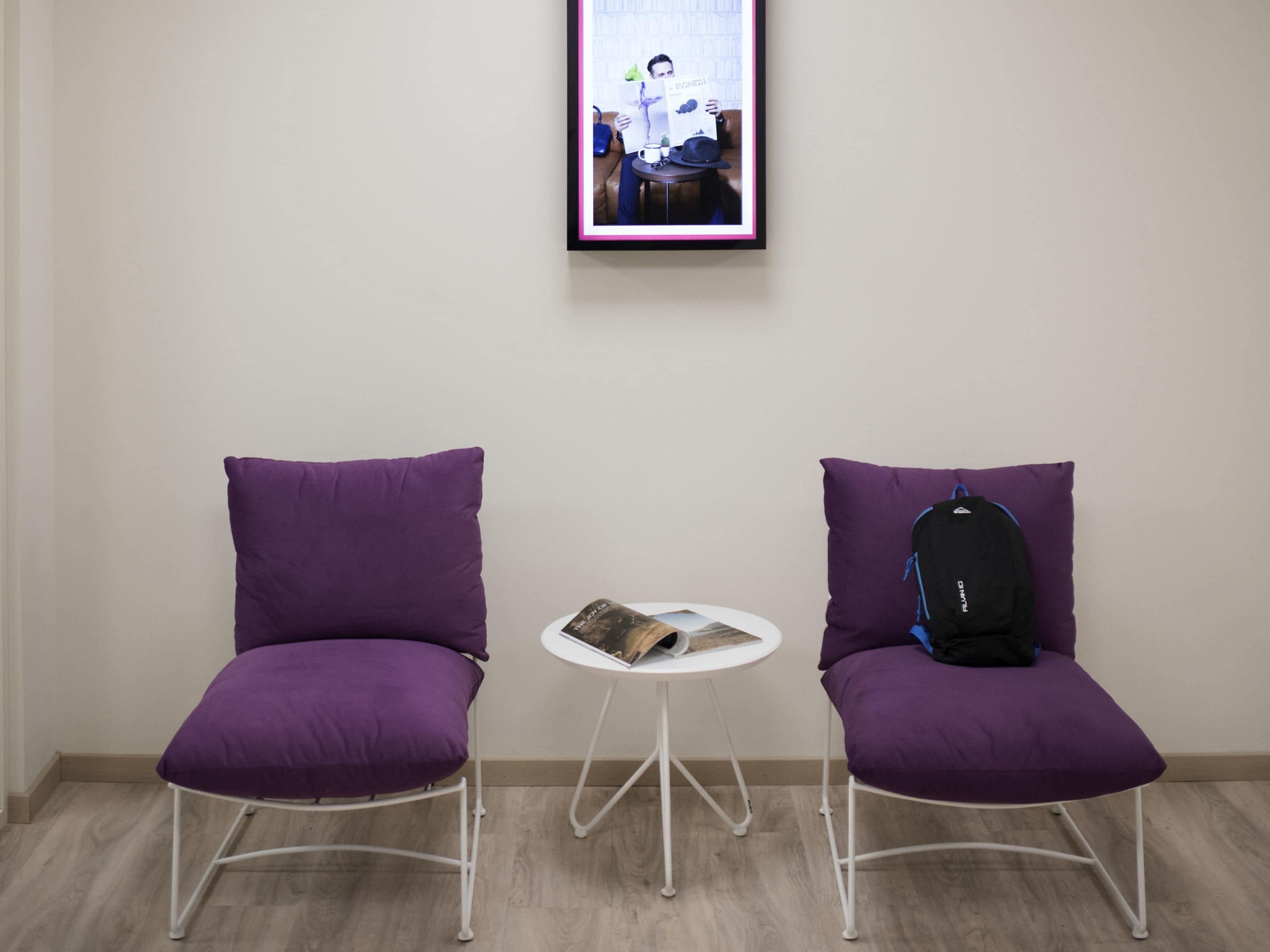 Zwei violette Stühle mit einem weißen Tisch dazwischen, ein gerahmtes Bild an der Wand und ein schwarzer Rucksack auf einem Stuhl.