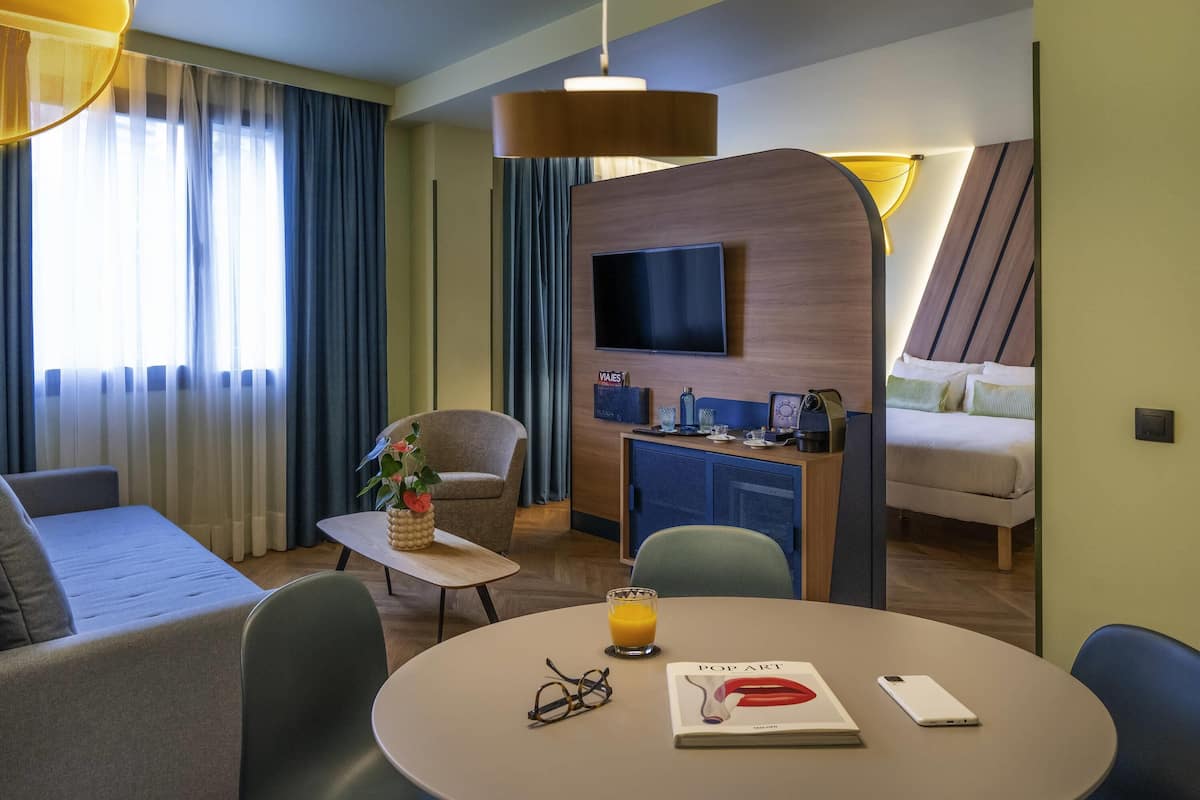 A hotel room with a sofa, a dining table with a book and glasses, a small table with a flower vase, a TV, and a bed.