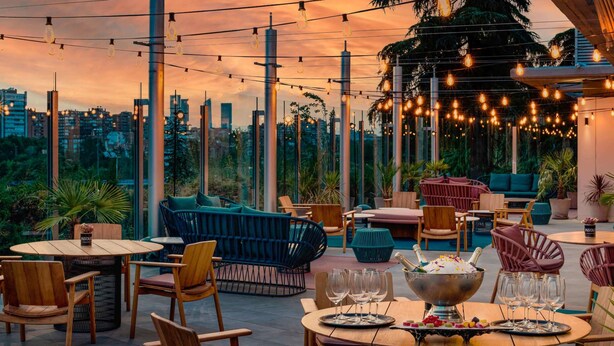 An outdoor seating area with tables and chairs, a large bowl with glasses and a bottle, and a cityscape view in the background.