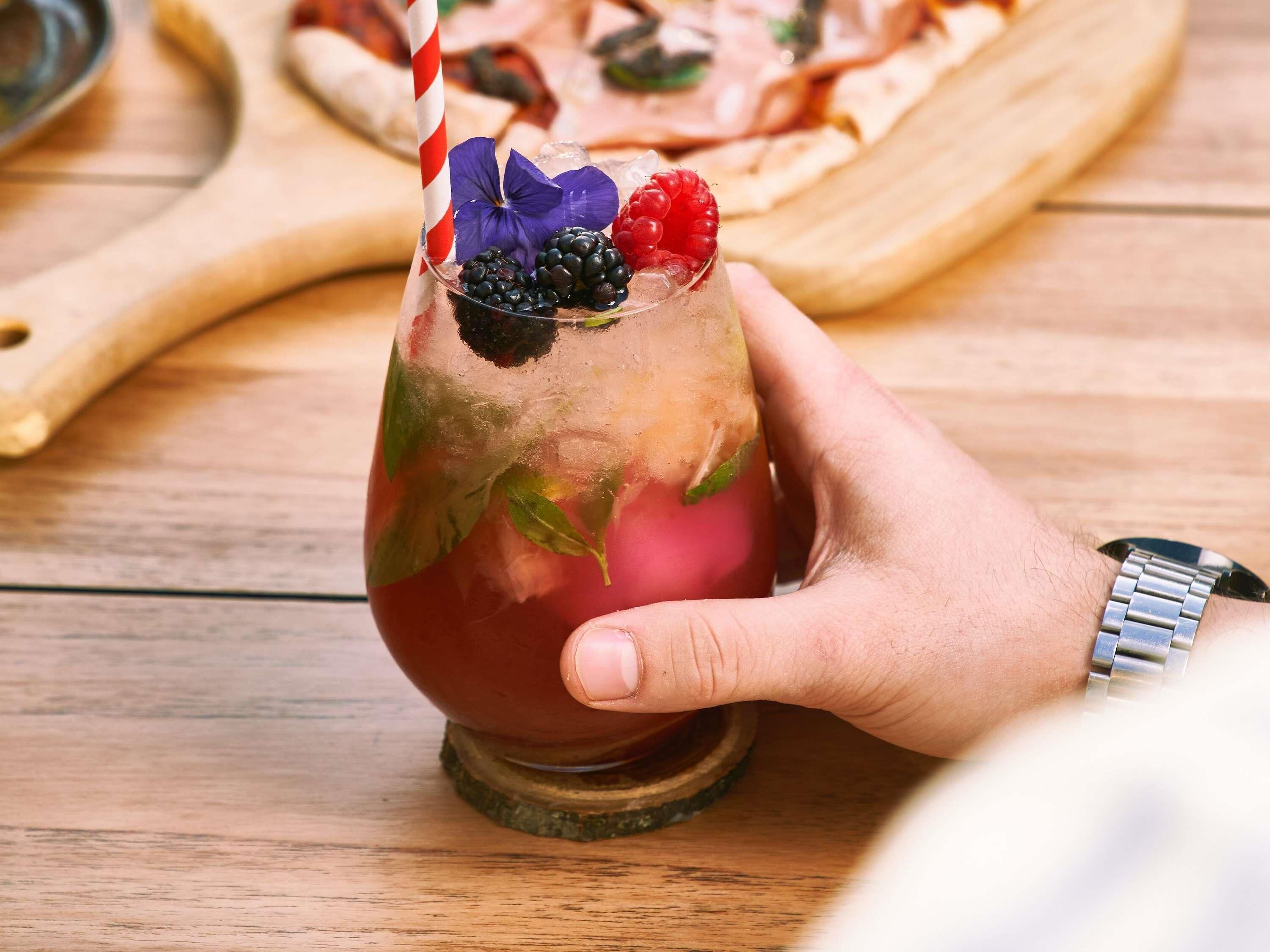 A hand holding a cocktail glass with a straw, garnished with berries and a flower, on a wooden table.