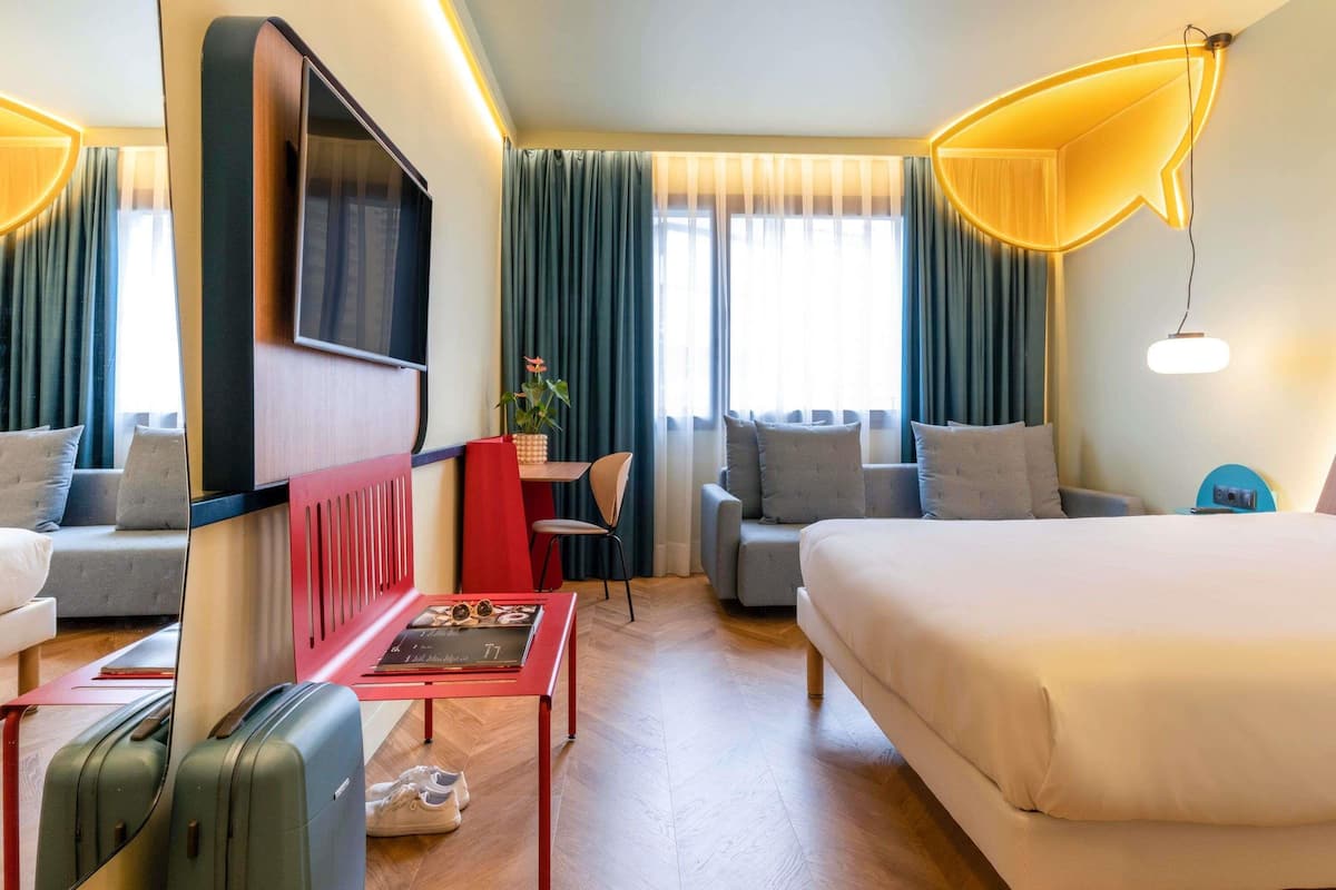 A hotel room with a bed, a desk, a chair, a TV, and a suitcase.