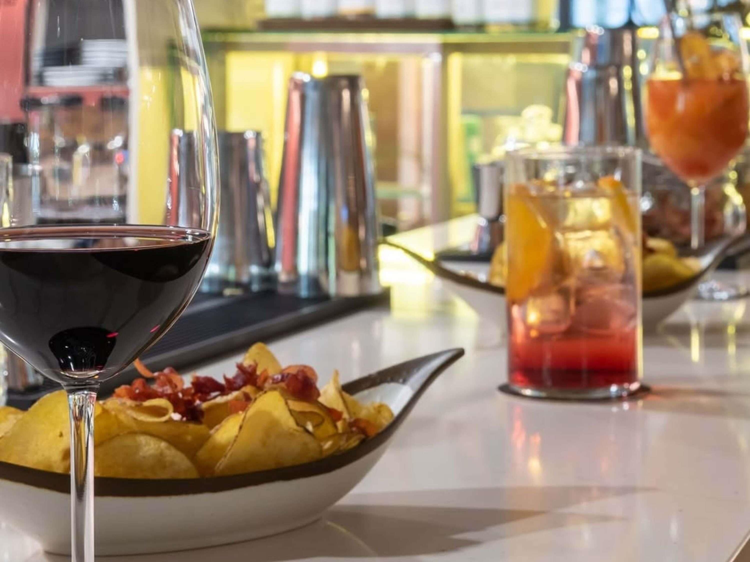 Una zona de bar con un bol de nachos, una copa de vino tinto y un cóctel con una rodaja de naranja.