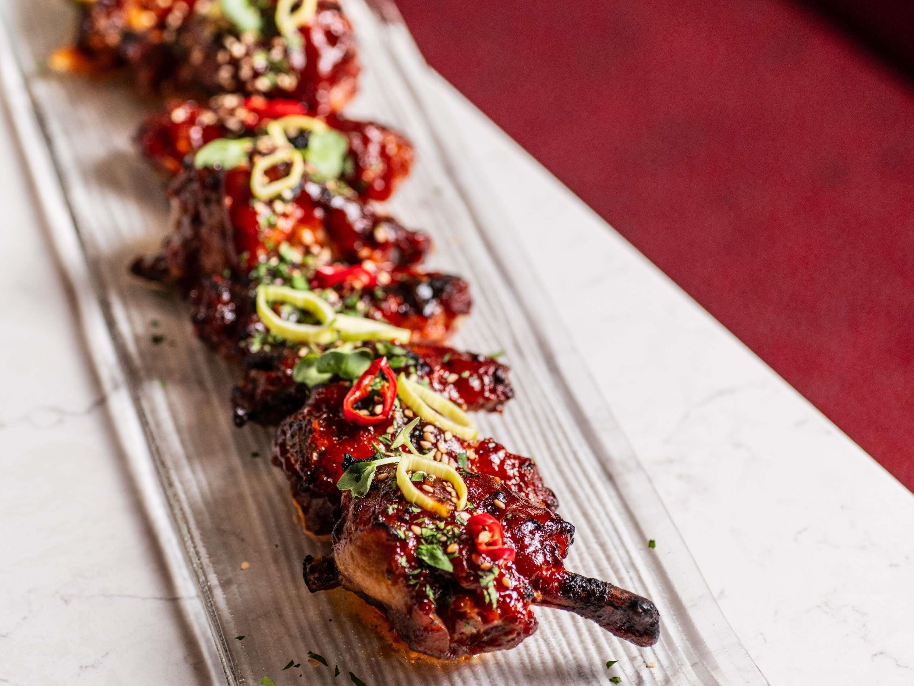 Une rangée de brochettes de viande, garnies d’herbes et de poivrons, est présentée sur une surface en marbre blanc.
