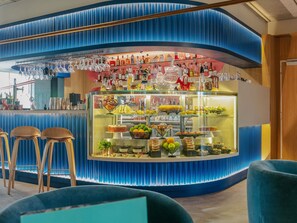 A bar area with stools, a glass display case with food items, and a variety of bottles. - Ibis Styles Madrid City las Ventas (Madrid)