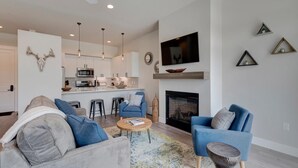 A modern living room with a grey sofa, blue armchairs, and a coffee table. A kitchen with white cabinets and a fireplace is visible in the background. - Downtown WP Loft 17 I Huge Hot Tub I Views I Bunk Beds I Game Console I Discounted Attractions (Winter Park)