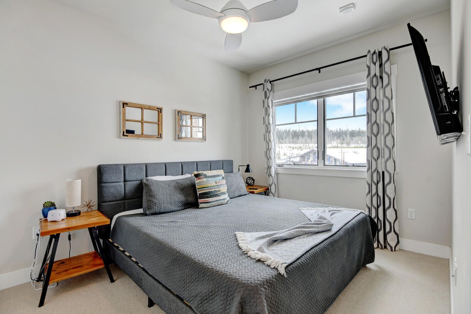 A bedroom with a large bed, a bedside table, a nightstand, a ceiling fan, a window with curtains, and a wall-mounted TV.