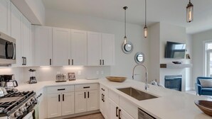 A modern kitchen with white cabinets, a stainless steel stove, and a dishwasher. There are pendant lights, a large island with a sink, and a flat-screen TV mounted above the fireplace. - Downtown WP Loft 17 I Huge Hot Tub I Views I Bunk Beds I Game Console I Discounted Attractions (Winter Park)