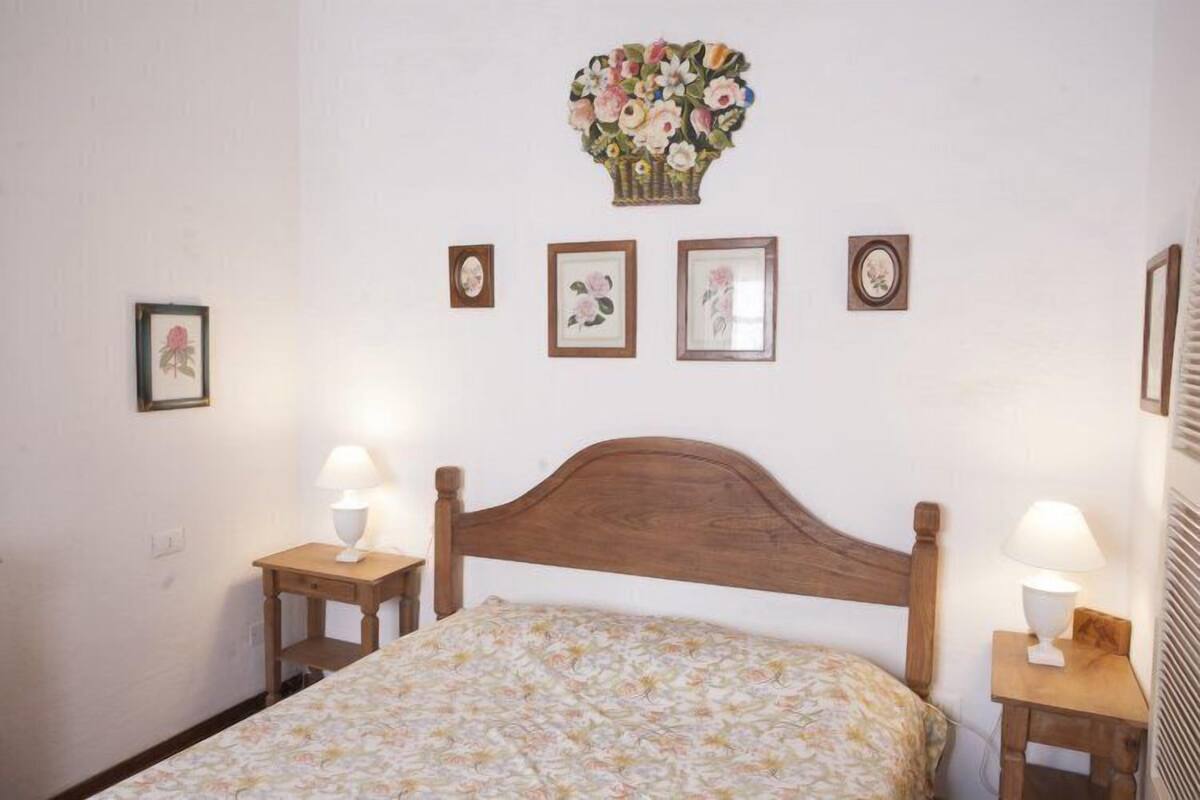 A bedroom with a wooden headboard, floral bedspread, two bedside tables with lamps, and four framed pictures on the wall.