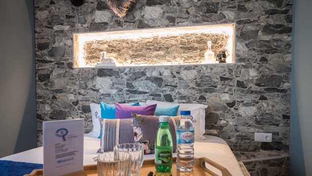 Un vassoio di legno con un menu, bicchieri, una bottiglia di Vimeiro e una di acqua, su un letto con una parete in pietra e una mensola illuminata sopra.