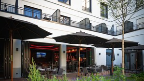 An outdoor seating area with umbrellas, tables, and chairs, and a building entrance with a bar visible inside. - Qube Hotel Bahnstadt (Heidelberg)