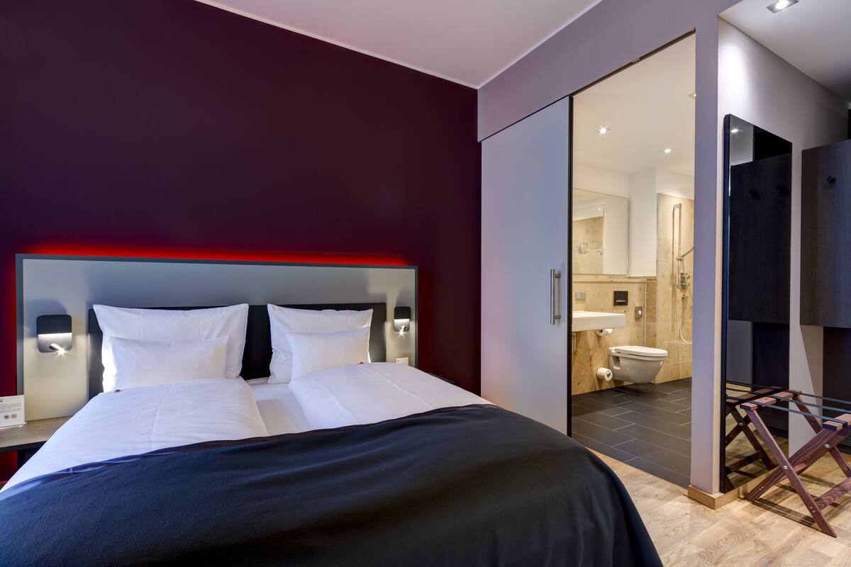 A modern hotel room with a large bed, a glass door leading to a bathroom, and a red accent on the headboard.