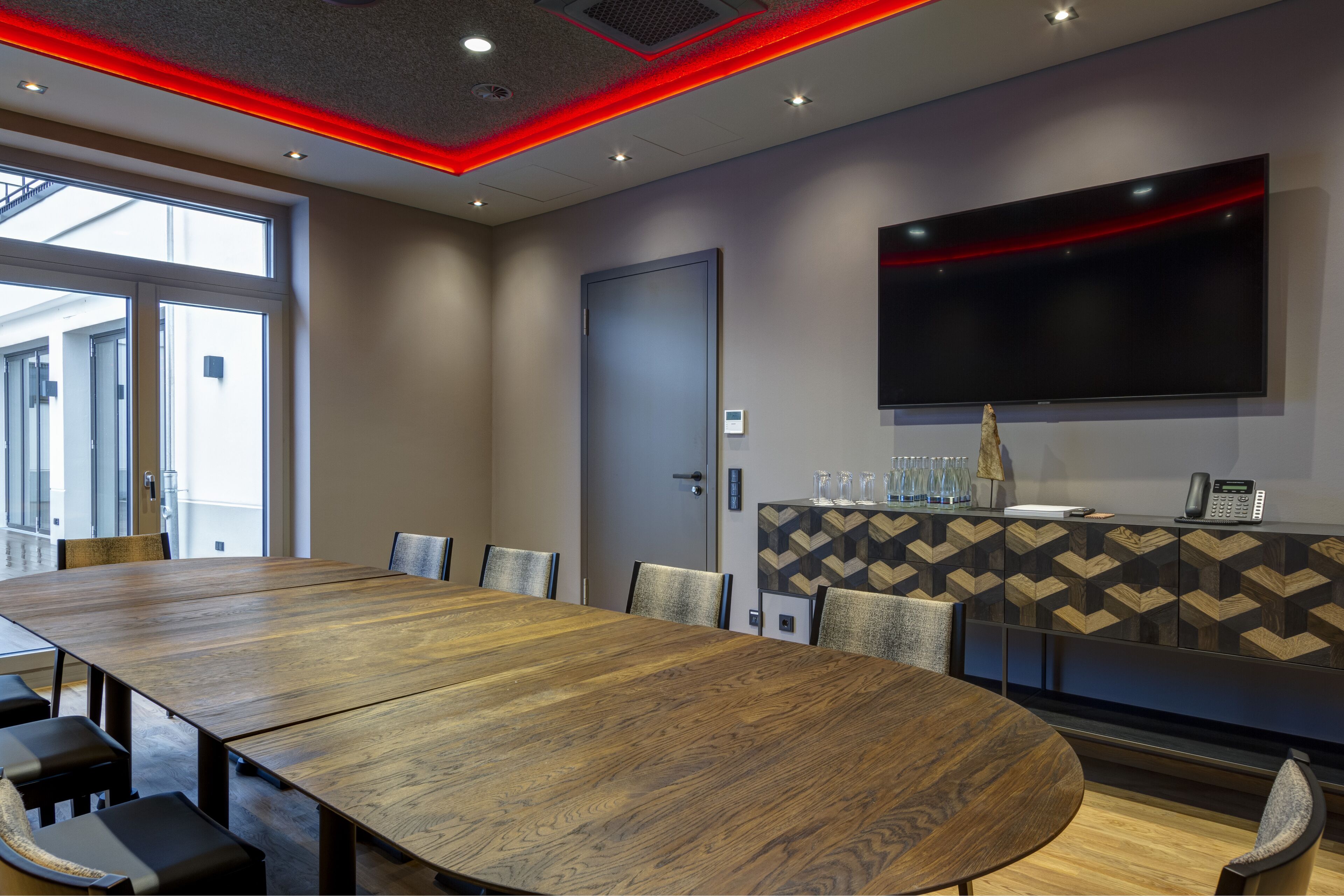 Uma sala de conferências com uma grande mesa de madeira, cadeiras, uma TV fixada na parede e um armário com padrão.