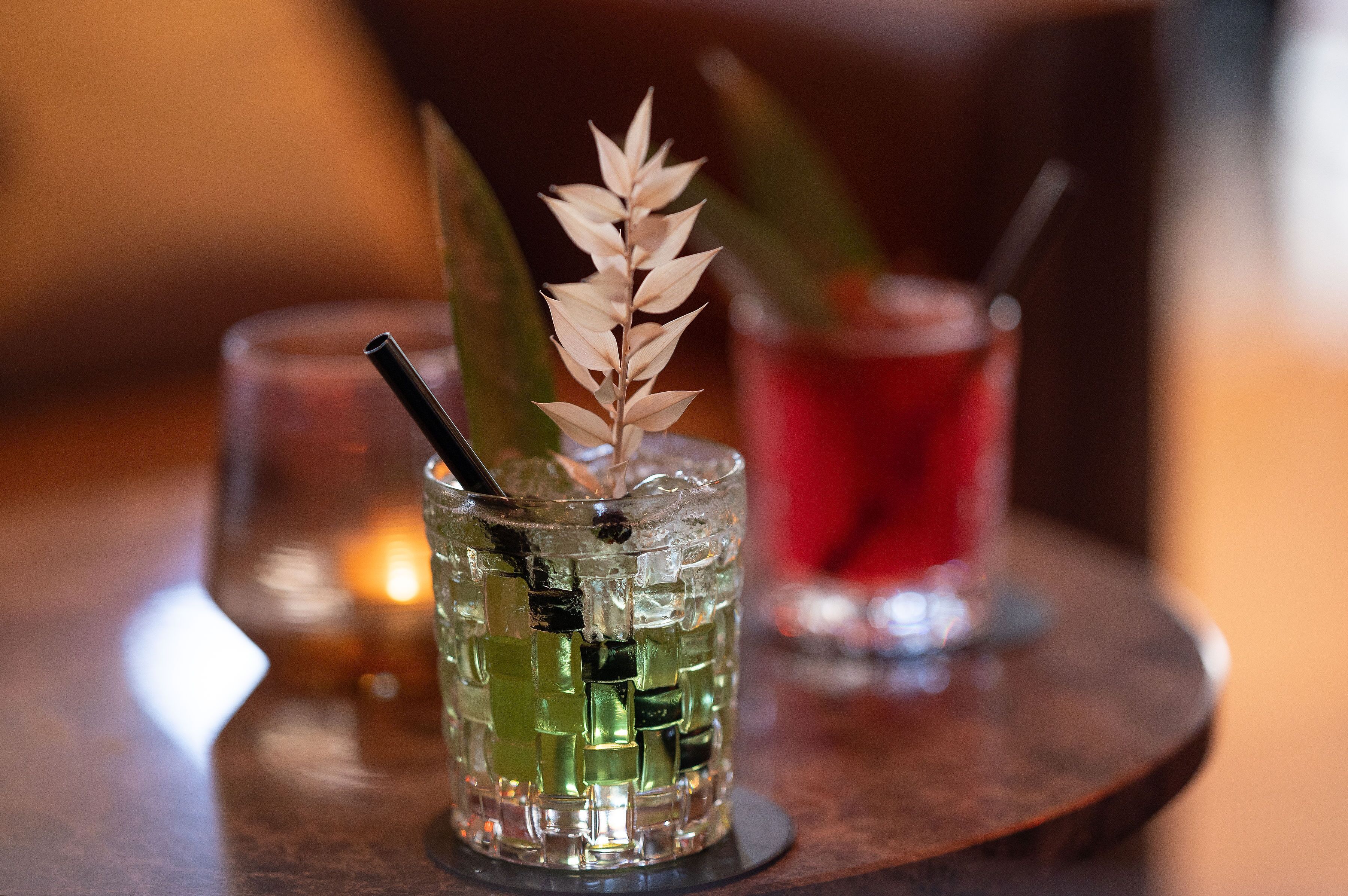 A glass with a decorative plant stem and a straw, placed on a wooden table with a candle in the background.