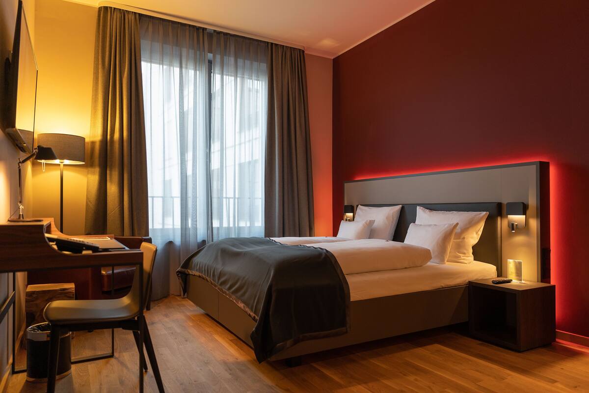 A hotel room with a bed, desk, chair, and a nightstand with a lamp.