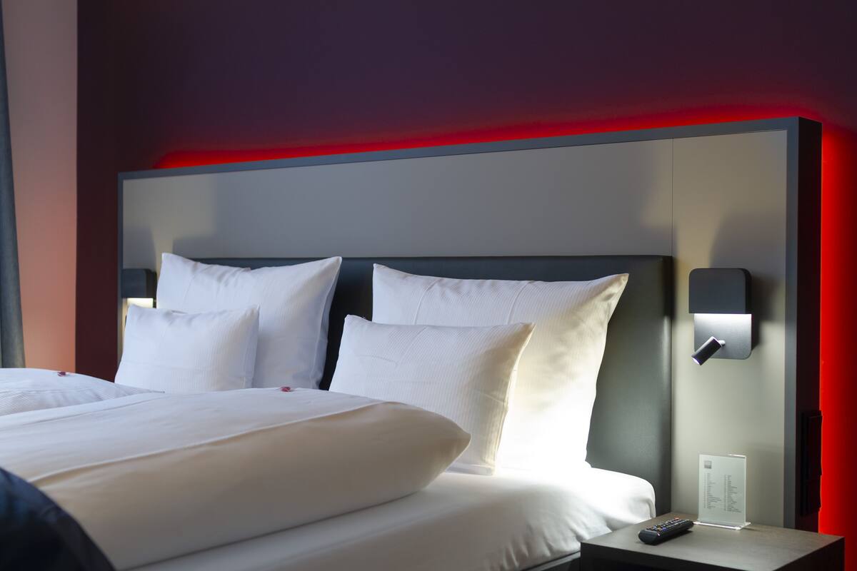 A hotel room with a large bed, two pillows, a nightstand with a remote control, and a wall-mounted light.