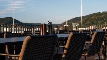 A restaurant deck with wicker chairs and tables, overlooking a body of water and hills.