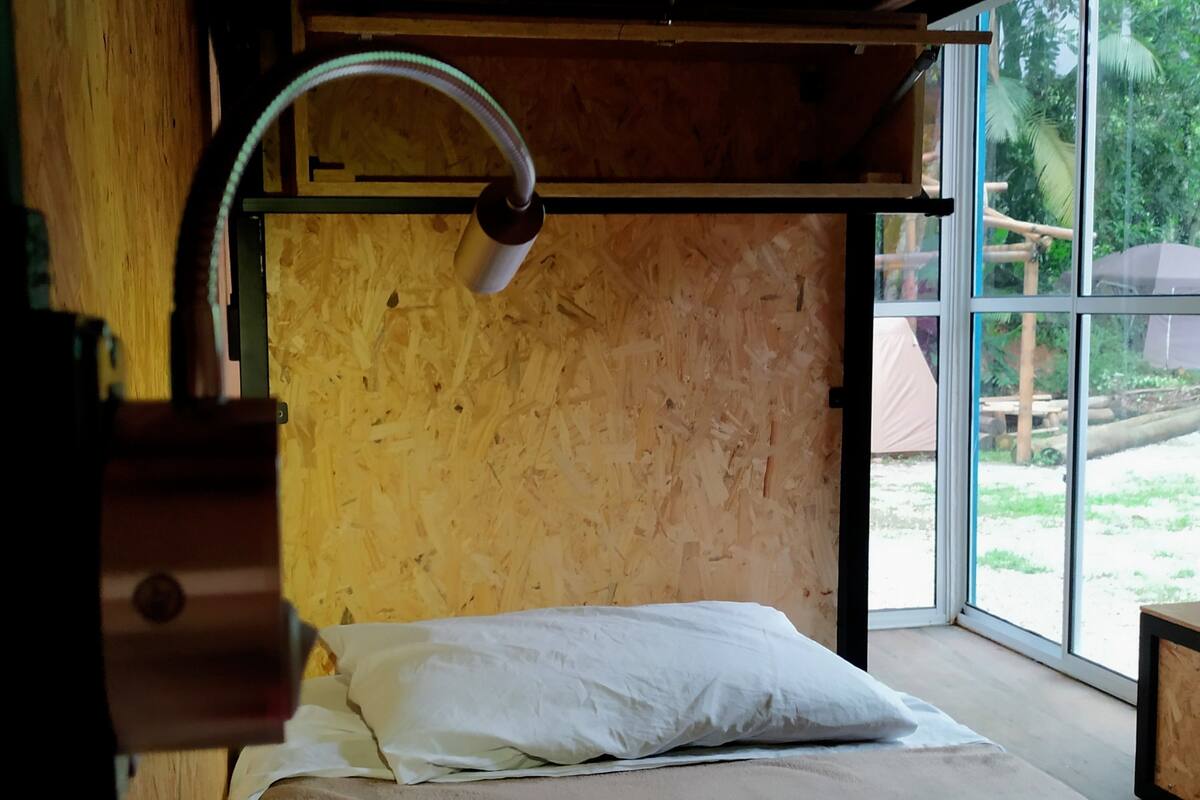 A room with a wooden wall, a bed with a white pillow, and a window providing a view of greenery outside.
